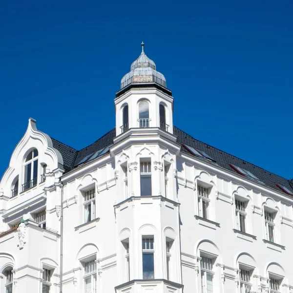 Weißes historisches Gebäude mit verzierten Fenstern und einem kleinen Turm unter einem klaren blauen Himmel, der an klassische Architektur erinnert - weitere Einzelheiten zu Ihrem Besuch finden Sie in unserer Datenschutzerklärung.