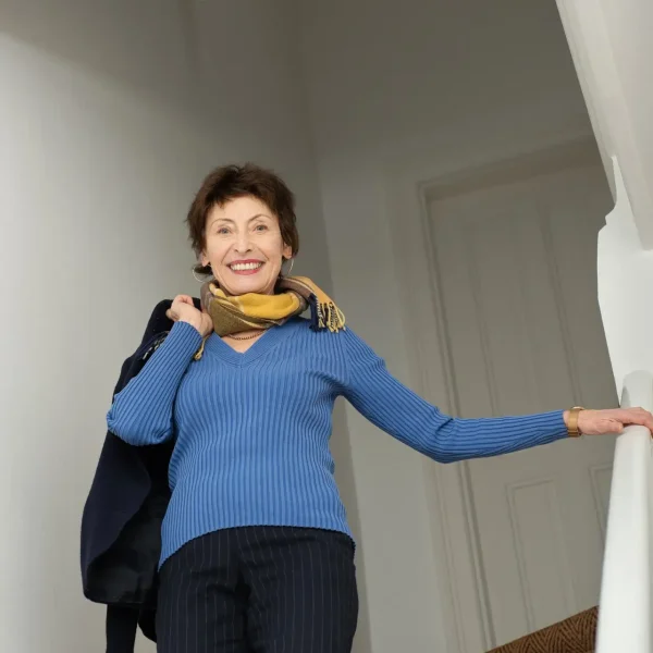 Eine lächelnde Frau in blauem Pullover und Schal geht eine mit Teppich ausgelegte Treppe hinunter und hält sich am Geländer fest, während in der Nähe ein Schild mit der Aufschrift "Datenschutzerklärung" erscheint.