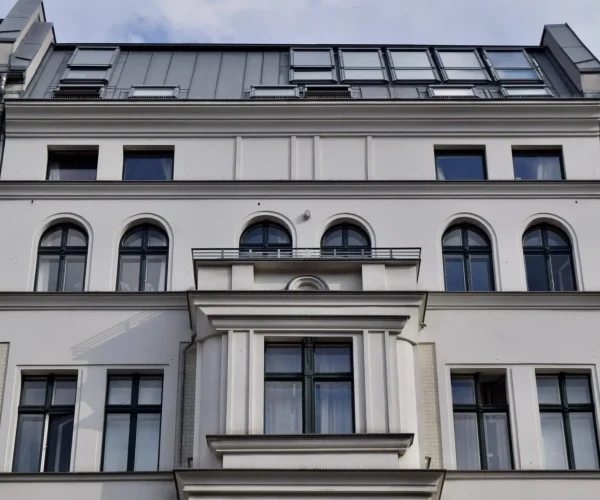 Weißes mehrstöckiges Gebäude mit bogenförmigen und rechteckigen Fenstern, einem modernen grauen Dach und einem klaren Blick unter einem bewölkten Himmel - Details finden Sie in unserer Datenschutzerklärung.