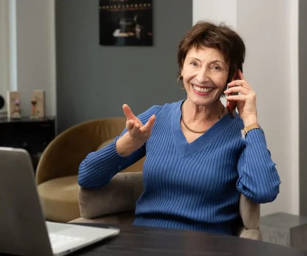 Lächelnde ältere Frau im blauen Pullover telefoniert am Schreibtisch mit Laptop in einem hellen, modernen Raum, vielleicht bespricht sie eine Datenschutzerklärung.