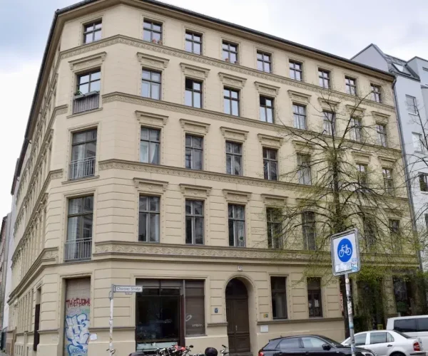 Beigefarbenes vierstöckiges Eckgebäude mit vielen Fenstern, geparkten Autos, Fahrrädern, einem blauen Radwegschild vor der Tür und einem sichtbaren Hinweis auf die Datenschutzerklärung in der Nähe des Eingangs.